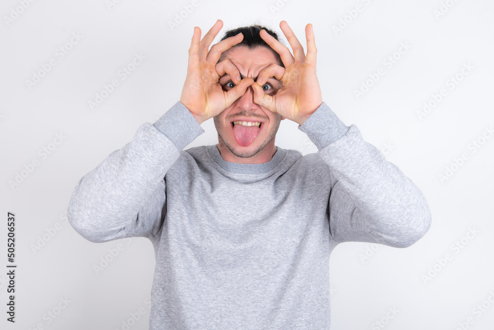 Young handsome dark haired man wearing fitted T-shirt over white wall doing ok gesture like binoculars sticking tongue out, eyes looking through fingers. Crazy expression. Young handsome dark haired man wearing fitted T-shirt over white wall doing ok gesture like binoculars sticking tongue out, eyes looking through fingers. Crazy expression.