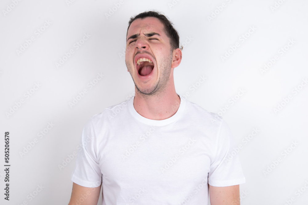 Young handsome dark haired man wearing fitted T-shirt over white wall angry and mad screaming frustrated and furious, shouting with anger. Rage and aggressive concept. Young handsome dark haired man wearing fitted T-shirt over white wall angry and mad screaming frustrated and furious, shouting with anger. Rage and aggressive concept.