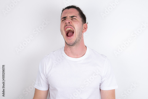 Young handsome dark haired man wearing fitted T-shirt over white wall angry and mad screaming frustrated and furious, shouting with anger. Rage and aggressive concept.