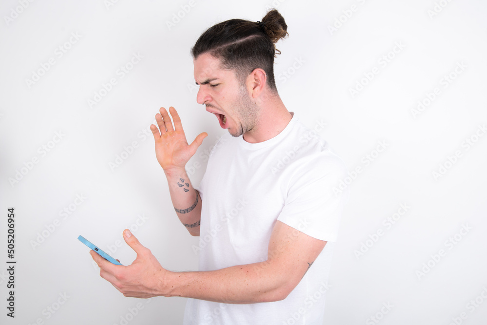 Angry Young handsome dark haired man wearing fitted T-shirt over white wall screaming on the phone, having an argument with an employee. Troubles at work. Angry Young handsome dark haired man wearing fitted T-shirt over white wall screaming on the phone, having an argument with an employee. Troubles at work.