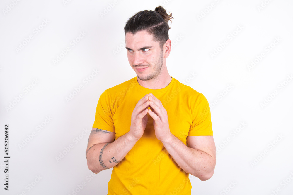 Young handsome dark haired man wearing fitted T-shirt over white wall steepled fingers and looks mysterious aside has great evil plan in mind