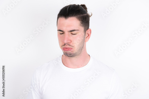 Dismal gloomy rejected Young handsome dark haired man wearing fitted T-shirt over white wall has problems and difficulties, curves lower lip and closes eyes in despair, being in depression
