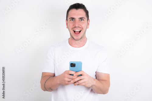 Excited Young handsome dark haired man wearing fitted T-shirt over white wall holding smartphone and looking amazed to the camera after receiving good news. © Roquillo