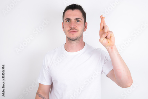 Young handsome dark haired man wearing fitted T-shirt over white wall pointing up with fingers number ten in Chinese sign language Shi