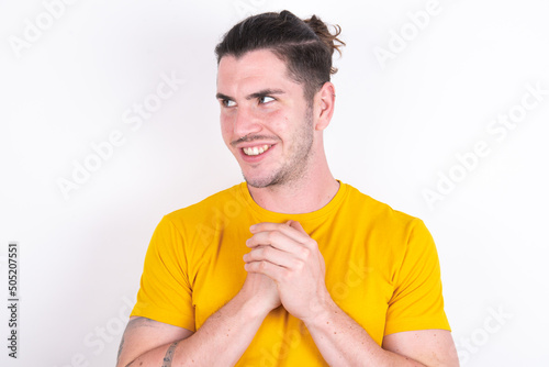 Happy young dark haired man wearing yellow t-shirt over white background anticipates something awesome happen, looks happily aside, keeps hands together near face, has glad expression.