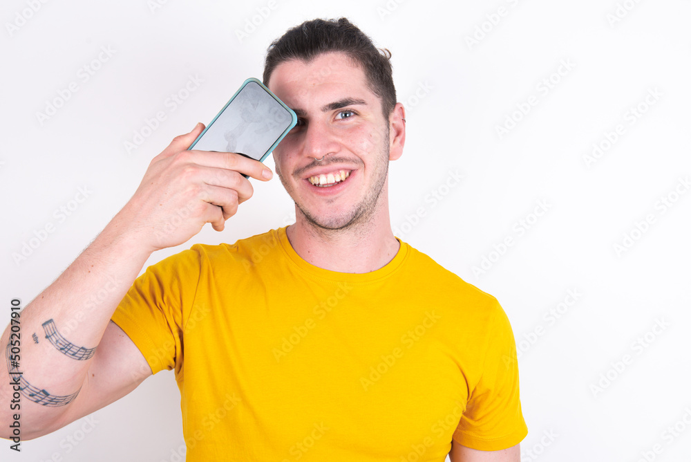 Young caucasian man wearing yellow t-shirt over white background holding modern smartphone covering one eye while smiling Young caucasian man wearing yellow t-shirt over white background holding modern smartphone covering one eye while smiling