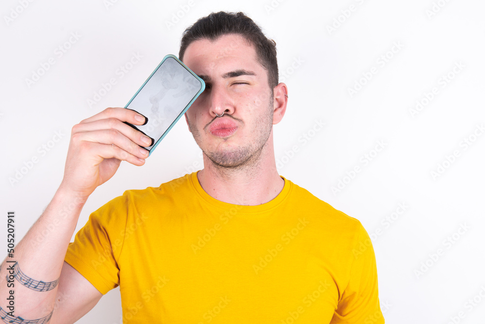 Adorable Young caucasian man wearing yellow t-shirt over white background holding modern device covering eye with lips pouted Adorable Young caucasian man wearing yellow t-shirt over white background holding modern device covering eye with lips pouted