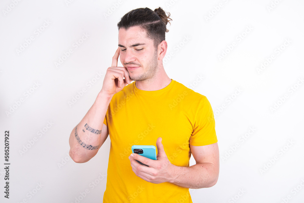 Young caucasian man wearing yellow t-shirt over white background holding gadget while sticking out tongue Young caucasian man wearing yellow t-shirt over white background holding gadget while sticking out tongue