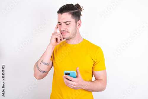 Young caucasian man wearing yellow t-shirt over white background holding gadget while sticking out tongue