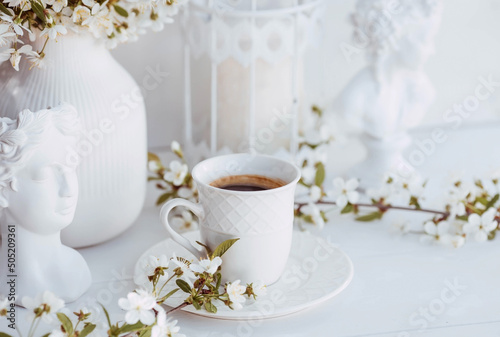 Fototapeta Naklejka Na Ścianę i Meble -  A beautiful postcard. A white coffee cup with a saucer, a statuette, candles and a vase with a bouquet of cherry blossoms. Beautiful still life. Spring time. The concept of 