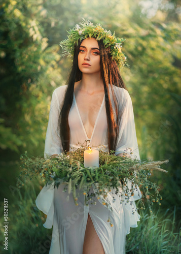 Portrait Fantasy Slavic woman holding herbal wreath, burning candle in hands. Divination ritual romantic girl witch sorceress conjures pagan ritual. White silk dress, long black hair. summer nature photo