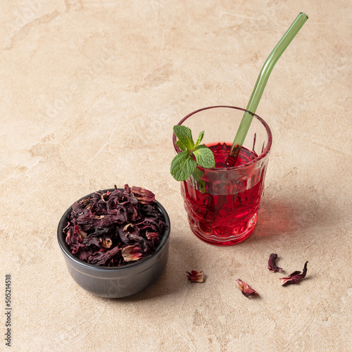 Karkade tea (hibiscus) in a glass with a glass tube for drinks and mint leaves. There is a ceramic bowl with dry tea nearby.