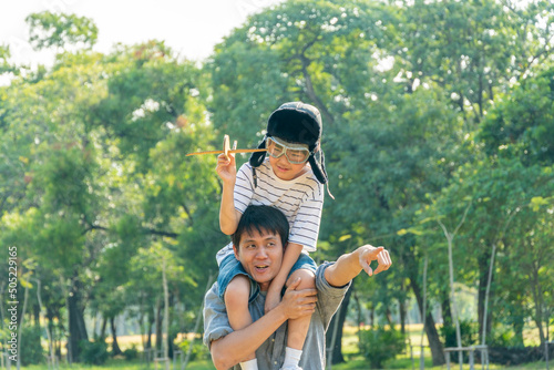 Asian boy wearing pilot helmet playing airplane toy sitting on father shoulder at park. dad support kid dream goal or aspiration. life living and family insurance concept. copy space