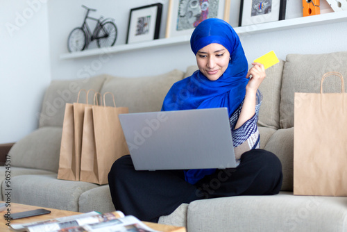 Young Muslim woman shopping online from home with credit card and laptop.