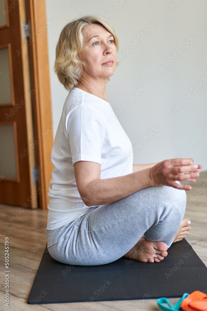 Sporty 60 year old gray haired woman sitting barefooted on yoga mat indoors doing bound ngle pose which helping to relieve symptoms of menopause. Aging, maturity, wellness and health