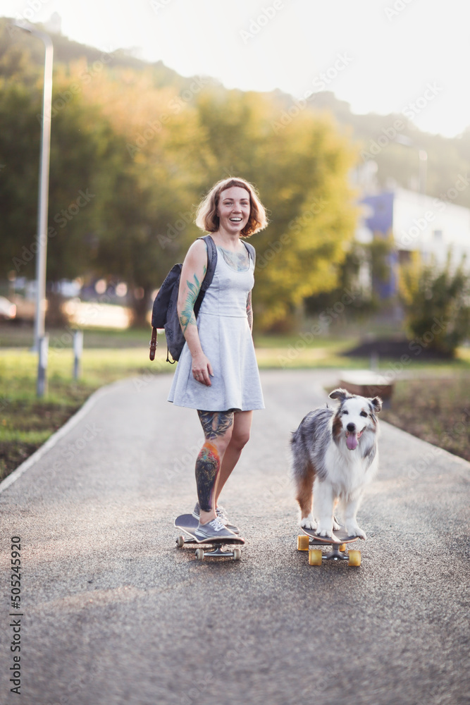 beautiful unusual girl with tattoos teaches and trains an Australian Shepherd dog to ride skateboard