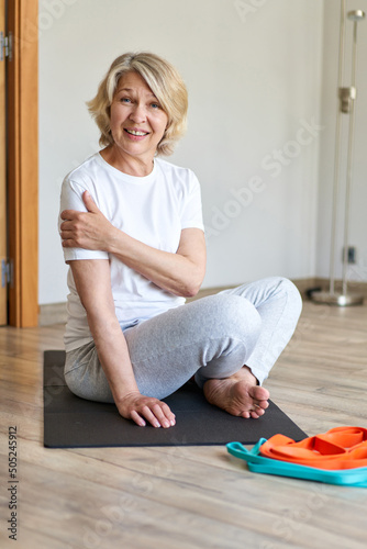 Sporty 60 year old gray haired woman sitting barefooted on yoga mat indoors doing bound ngle pose which helping to relieve symptoms of menopause. Aging, maturity, wellness and health