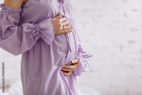 Side view close up cropped of pregnant woman in purple dress touching belly tummy, expecting baby, young future mother standing at home on white brick background, copy space, motherhood concept photo