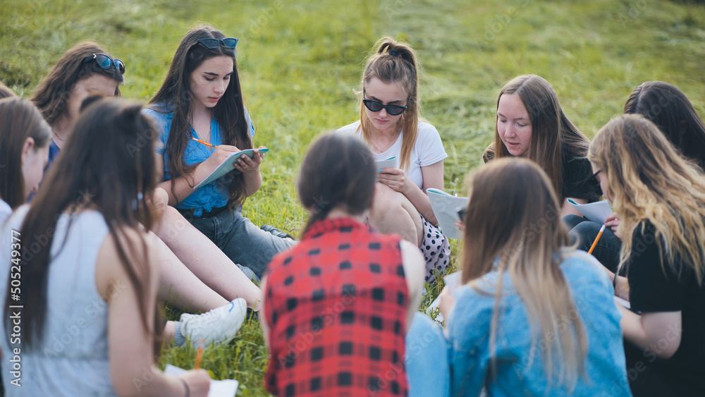 A group of female students are sitting in a circle on a meadow for collective work with notebooks. A group of female students are sitting in a circle on a meadow for collective work with notebooks.