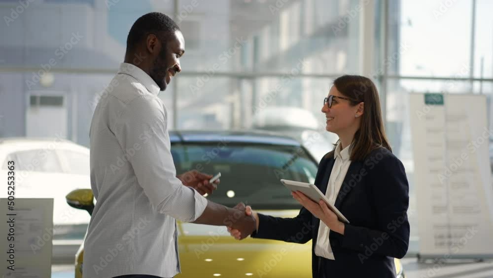 Woman manager is giving car keys to male customer and standing in dealership office spbas. 4k Side view of beautiful caucasian woman gives keys to African man and looks with smile, makes handshake and