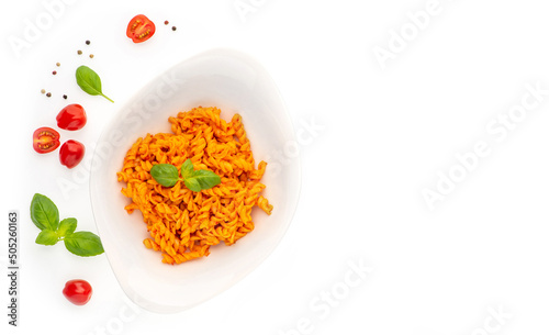 Flatlay composition of fusilli pasta with paprika pesto sauce and basil leafs in porcelain plate and cherry tomatoes, and pepper grains around on white isolated background