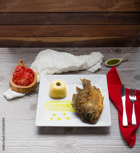 Fried fish accompanied by mashed potatoes with coriander oil and fresh tomato, served on a square white plate.