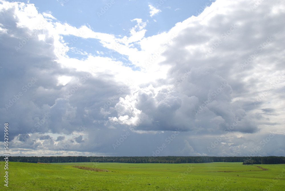 White-blue clouds over a green field. Above a large uneven field with green grass along the edges of which a forest grows, large white rain clouds hang low through which the blue sky peeps through. White-blue clouds over a green field. Above a large uneven field with green grass along the edges of which a forest grows, large white rain clouds hang low through which the blue sky peeps through.