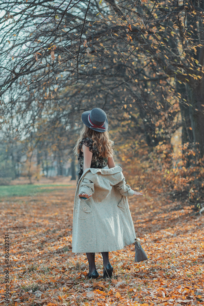 happy young curly girl dressed in a white coat and hat in a thin dress and heels with a red handbag on her shoulders, the girl spins in the middle of yellow fallen leaves in the autumn park in sunn