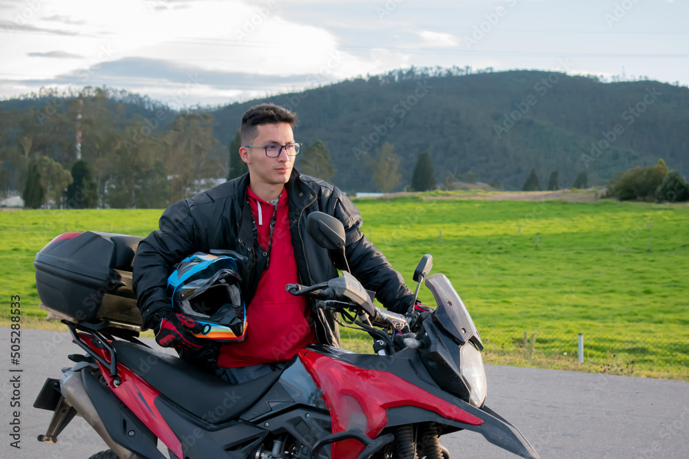 Obraz premium hombre parado al lado de una moto con un casco azul en la mano y fondo de montañas 