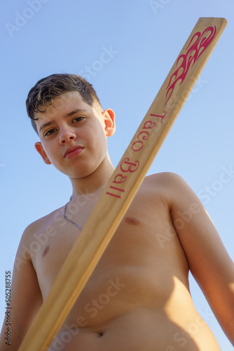 garoto com expressão seria segurando taco de madeira na mão, sem camisa, na praia. Jogo de taco, semelhante ao baisebol. photo