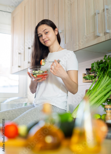 Young housewoman eating vegetable salad from plate at home kitchen photo
