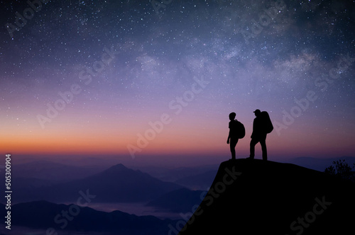 Silhouette of two young traveler with backpack standing on top of the mountain watched the beautiful view of night sky, star and milky way. We are happy to travel with friend in the wide world.