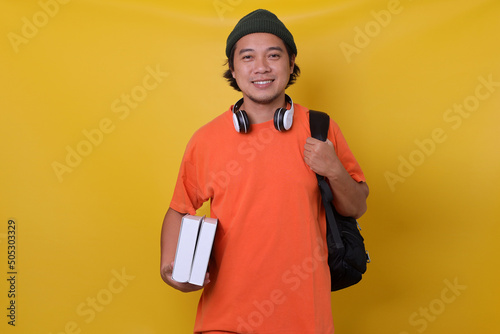 Fotografie Asian young man in casual style with backpack and headphone bringing books isolated on yellow background