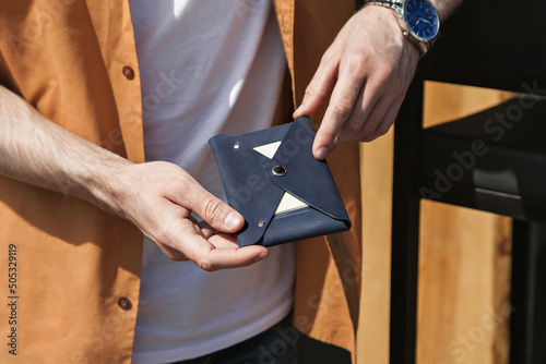 A man on the street in a white T-shirt and an orange shirt is holding a wallet with credit cards made of dark blue leather. Close-up, no face, copy space, horizontal orientation.