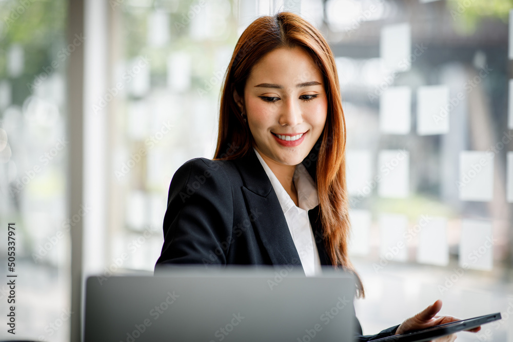 Photo of cheerful pretty Asian woman having been employed to a job as executive smiling toothily sitting at the desktop with laptop and writing note. Photo of cheerful pretty Asian woman having been employed to a job as executive smiling toothily sitting at the desktop with laptop and writing note.