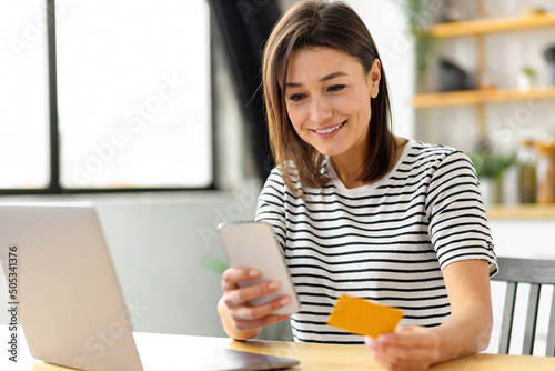 Online shopping, technology concept. Happy Caucasian woman holding mobile phone and credit card sitting at the table in the home kitchen