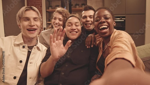 Handheld POV slowmo of five young cheerful LGBTQ friends smiling and waving at camera sitting on sofa together having cozy home party in modern loft style apartment photo