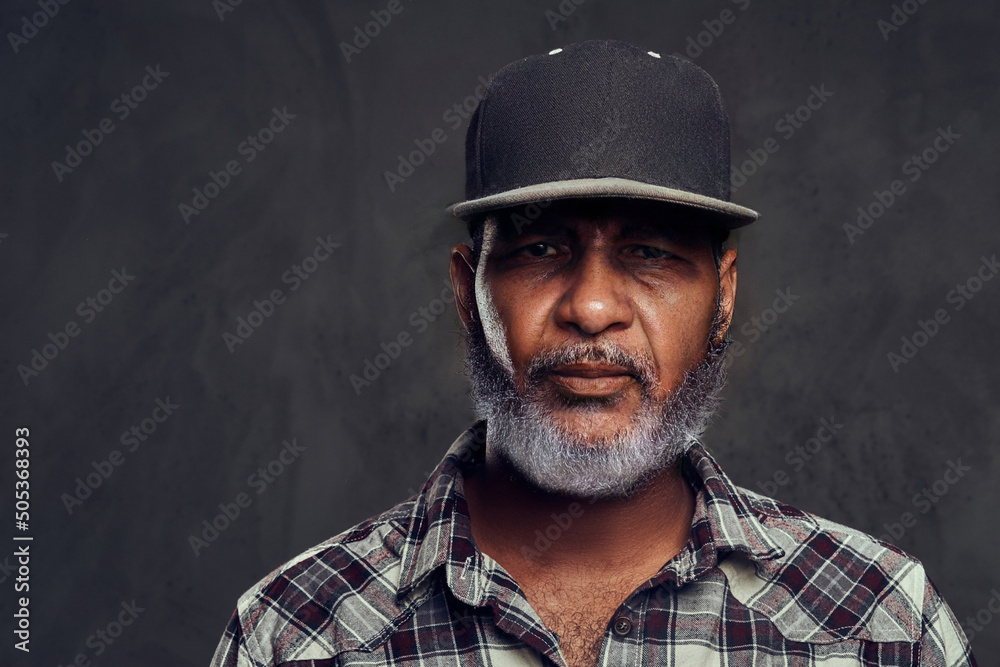 Shot of senior man dressed in plaid shirt and cap looking at camera against dark background. Shot of senior man dressed in plaid shirt and cap looking at camera against dark background.
