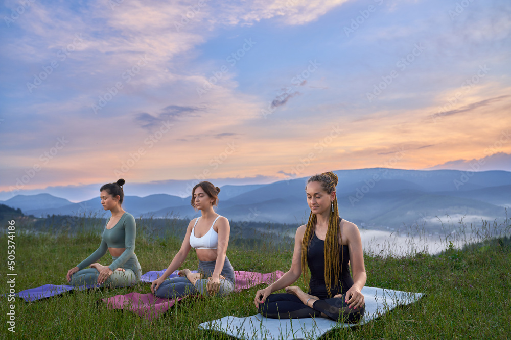 Three beautiful women with closed eyes meditating on yoga mat in lotus pose, deeply breathing and smiling. Pretty female among grass relaxing after yoga practice at sunrise time. Concept of balance. Three beautiful women with closed eyes meditating on yoga mat in lotus pose, deeply breathing and smiling. Pretty female among grass relaxing after yoga practice at sunrise time. Concept of balance.