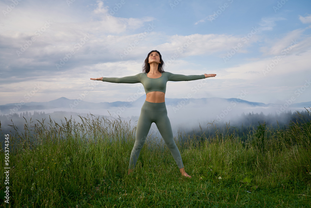 Front view of brunette woman standing on grass on mountain hill in pose of winner, looking up. Attractive female doing yoga outdoor in morning time among fog. Concept of health. Front view of brunette woman standing on grass on mountain hill in pose of winner, looking up. Attractive female doing yoga outdoor in morning time among fog. Concept of health.