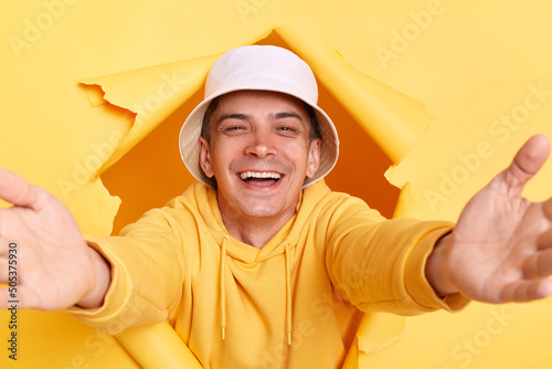 Portrait of kind pleased man wearing casual hoodie and panama looking through torn hole in yellow paper, giving free hugs with outstretched hands, welcoming inviting to embrace, support and care.