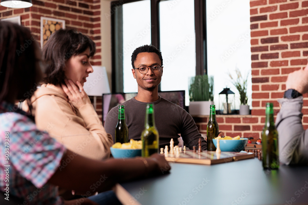 Smiling man sitting at table in living room with friends while watching a game of chess. Intelligent multiethnic people playing strategy boardgames while enjoying snacks and beverages. Smiling man sitting at table in living room with friends while watching a game of chess. Intelligent multiethnic people playing strategy boardgames while enjoying snacks and beverages.