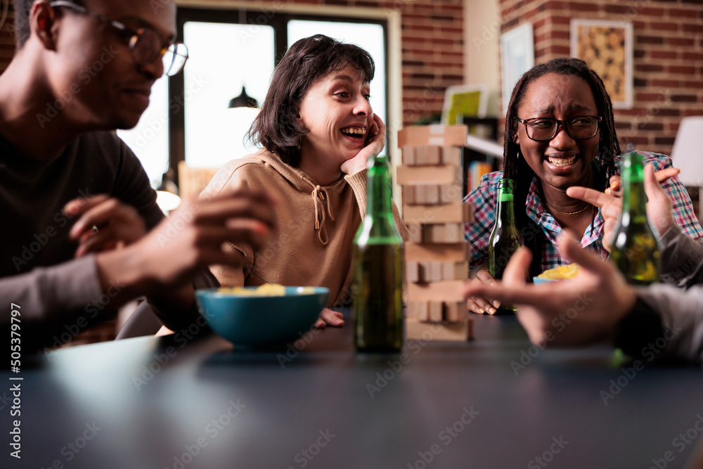 Diverse group of friends laughing together while discussing and playing society games at home. Multiethnic people sitting at table in living room while enjoying fun leisure activity with wood blocks.
