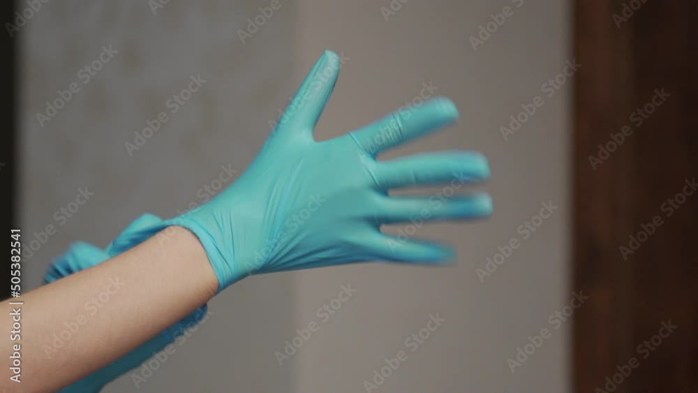 Young woman puts on blue rubber gloves on hands on blurred background in room. Housewife gets prepared for cleaning dust in apartment close view