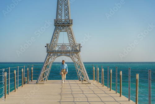Large model of the Eiffel Tower on the beach. A woman walks along the pier towards the tower, wearing a blue jacket and white jeans.