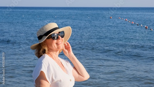 beautiful 50 year old woman in a straw hat and sunglasses on a blue sea background. Summer, vacation, vacation, active retirees