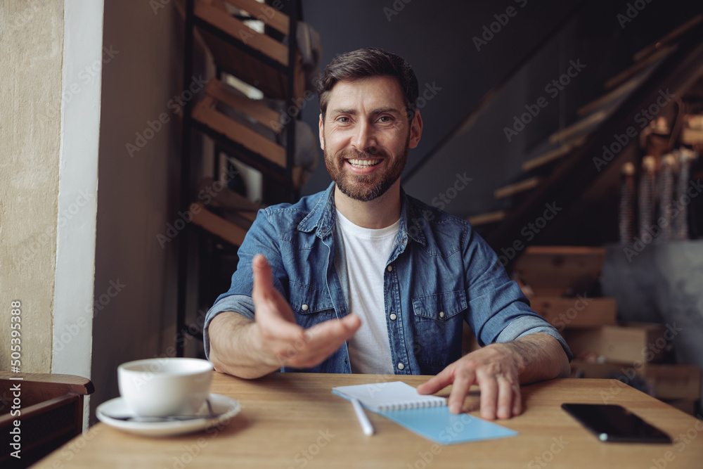 Joyful attractive man reaching for camera with hand. Coffee time in cafe. Portrait. Give me gesture.