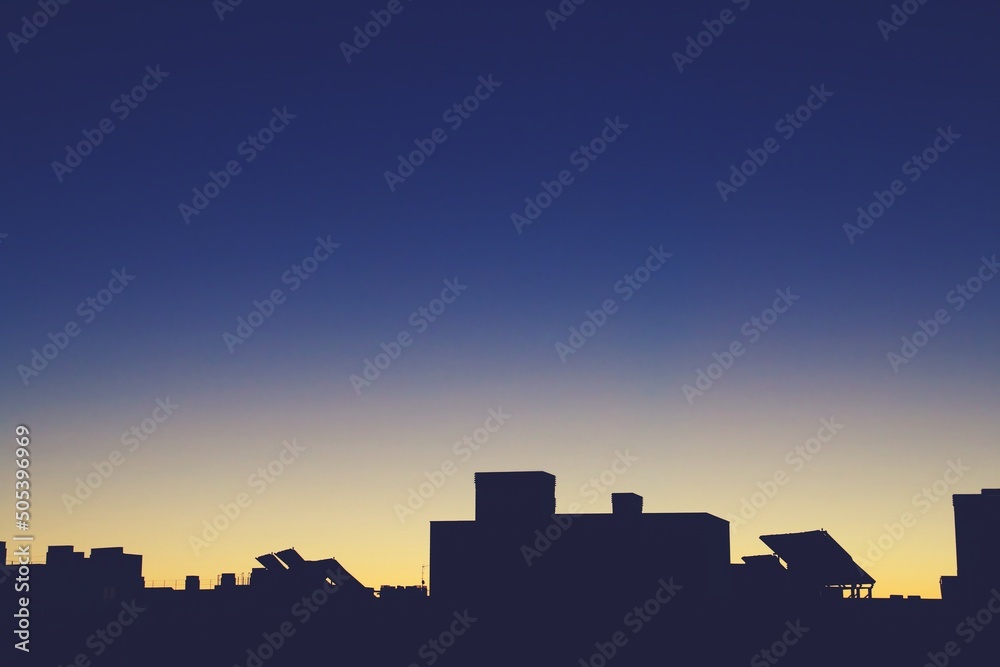 Silueta de la parte superior de un edificio moderno en Madrid, España. Silueta de las placas solares y chimeneas situadas en la azotea con los primeros rayos de sol en un cielo amarillo y azul.