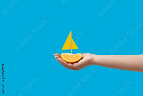 Summer concept, hand holding sailing boat made of orange on pastel blue background. Minimal tropical creative concept.