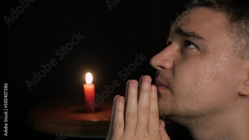 Close-up of young caucasian male praying to God with yellow candle in background. Sinner folding hands together, looking up and saying a quiet prayer to Jesus, begging for forgiveness for his soul. 
